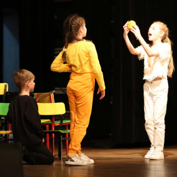 Czworo dzieci na scenie. Dwoje klęczy na podłodze. Dwie dziwczynki stoją naprzeciw siebie. Jedna trzyma wysoko w dłoniach żółty przedmiot i szeroko otwiera usta.