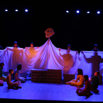 Pięć osób trzyma rozciagniętą wzdłuż białą tkaninę. W górze maska. Na scenie siedzi siedmioro dzieci. Mrok. Cztery punktowe światełka.