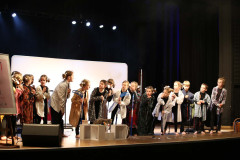Grupa dzieci na scenie podczas występu. Dziewczynki mają włosy związane w kitki. Wszystkie trzymają kije trekkingowe. Zdjęcie w planie ogólnym.