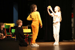 Czworo dzieci na scenie. Dwoje klęczy na podłodze. Dwie dziwczynki stoją naprzeciw siebie. Jedna trzyma wysoko w dłoniach żółty przedmiot i szeroko otwiera usta.