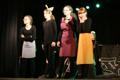 Cztery dziewczyny na brzegu sceny. Stoją w rzędzie odgrywając role. W tle ciemna kurtyna i fragment scenografii - na czarnym materialne namalowane białe zarysy naczyń kuchennych.