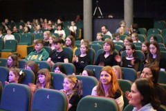 Dzieci siedzące na widowni w zielonych numerowanych fotelach kinowych.