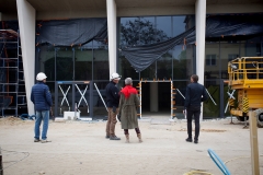 Stojąca tyłem do obiektywu grupa osób przed głównym wejściem do budynku. Szklane elemnty przykryte ciemną folią.