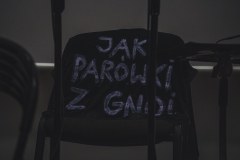 Przewieszona przez krzesło ciemna kosulka z białym napisem: JAK PARÓWKI Z GNOI.