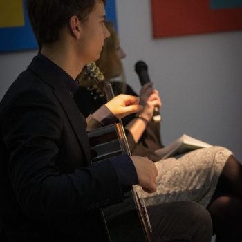 Sfotografowany z prawego profilu chłopak trzymający na kolanach gitarę. W głębi prowadząca spotkanie Małgorzata Siemianowska trzyma mikrofon.