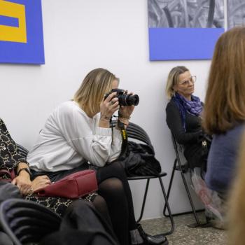 Siedzące przy ścianie trzy kobiety. Kobieta w środku robi zdjęcie aparatem.