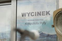 Fragment rozsuwanych drzwi budynku CKiS przy ul. Okólnej w Koninie z naklejonym plakatem reklamującym wystawę "Wycinek Krzysztofa Ryfy". W środku kadru nieostry mikrofon na statywie, z prawej strony fragment reflektora