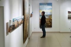 Wyeksponowane na białej ścianie obrazy Krzysztofa Ryfy, W głębi nieostra postać mężczyzny oglądającego jedną z prac.