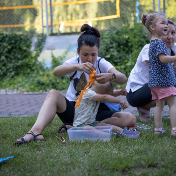 Na trawniku dwie kobiety i dzieci. Przed nimi plastikowe pudełka.