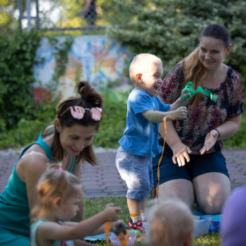 Kobiety z dziećmi. W centrum kadru chłopiec potrząsający kolorowymi paskami bibuły. W tle murek porośniętymi roślinami z fragmentami graffiti.