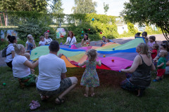 Na trawniku opiekunowie i dzieci tworzą koło. Trzymają między sobą tęczową chustę. Na chuście kolorowe piłki.