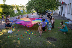 Kobiety z dziećmi trzymają tęczową chustę. Piłki leżą na trawniku. Po lewej murek z siatką obrośnięty bluszczem. Po prawej budynek. W głębi drzewo.