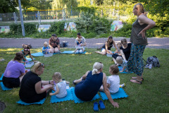 Kobiety z dziećmi siedzą na niebieskich matach w kształcie puzzli. Po prawej stoi prowadząca zajęcia.