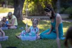 Na trawniku siedzą kobieta i dziewczynka. Dziecko trzyma drewniane szczypce do prania nad plastikowym pudełkiem.