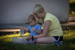 Siedząca na trawniku kobieta z dzieckiem. Chłopiec trzyma sitko nad plastikowym pudełkiem.