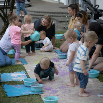 Na ubrudzonej farbami tekturze dzieci bawią się miskami. Wzdłuż maty poplamione farba siedziska w kształcie puzzli. Na trawie klęczą opiekunki.