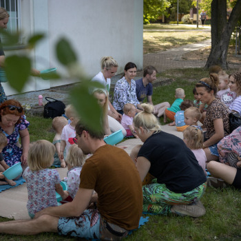 Zdjęcie grupowe. Rodzice z dziećmi siedzą z miskami przy falistej tekturze rozłożonej na trawie.