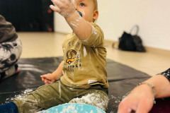 Oprószone mąką dziecko siedzi na czarnej folii. Podnosi w górę lewą rękę. Przed nimi plastikowa miska.