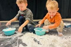 Dwóch chłopców przy miskach z mąką. Przed nimi wysypana mąka na czarnej folii. Dziecko po prawej w pomarańczowej bluzie, po lewej - w szarej. W tle ciemna kotara.