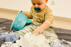 Dziecko trzyma dłoń nad babką usypaną z mąki. W prawej dłoni trzyma plastikową miskę. W buzi smoczek.