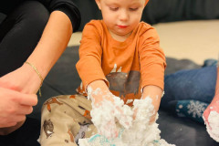 Dziecko siedzi na czarnej folii przy misce z białą mazią, do której wkłada dłonie. Ubrane w pomarańczową bluzkę. Po prawej fragment nogi, po lewej fragment przedramienia.