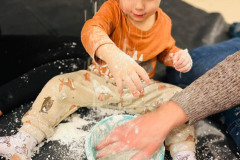 Dziecko siedzi okrakiem przy plastikowej misce z mąką. W misce dłoń.