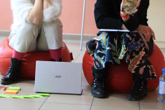 Dwie kobiety siedzą na czerwonych pufach. Przed nimi otwarty laptop, karteczki samoprzylepne i rozrzucone karty.