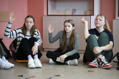 Trzy siedzące na podłodze dziewczyny podnoszą do góry prawe ręce. Za nimi szafka.