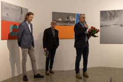 Na podwyższeniu stoją od lewej: Marcin Kochowicz, dyrektor MBWA w Lesznie, uśmiechający się do obiektywu Robert Brzęcki, kurator galerii oraz Sławomir Kuszczak z bukietem róż i mikrofonem. Zdjęcie w planie ogólnym.