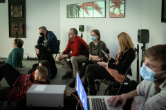 Grupa osób siedzących na plastikowych krzesłach lub półleżących na podłodze. W prawym dolnym rogu zamaskowany chłopak przy laptopie.