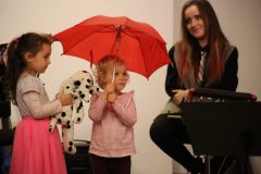 Z lewej strony dziewczynka z pluszowym dalmatyńczykiem. W środku dziewczynka trzyma czerwoną parasolkę. Po prawej członkini zespołu. Wszystkie są na scenie.