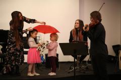 Dwie dziewczynki pod parasolem, który trzyma jedna z artystek. Mężczyzna gra na skrzypcach. Za nim na scenie siedzi jeszcze jedna członki zespołu.