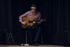 Siedzący na scenie mężczyzna gra na gitarze.