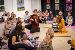 Na podłodze na rozłożonych kolorowych matach siedzą rodzice z dziećmi. Między nimi klęczący na jednym kolanie mężczyzna gra na skrzypcach.
