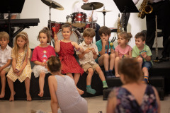 Dzieci siedzą w rzędzie na brzegu sceny. Za ich plecami widoczne instrumenty muzyczne.