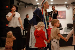 Uczestnicy zajęć w trakcie tańca z kobietą w stroju ludowym. Po lewo kobieta z mikrofonem i wpatrzone w nią dziecko. W głębi kadru muzycy na scenie.