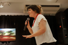 Sfotografowana z lewego profilu kobieta z mikrofonem. Ubrana w białą bluzkę. Na szyi czerwone korale.