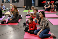 Uczestnicy zajęć: dorośli z dziećmi siedzą na kolorowych matach rozłożonych na szarej podłodze.