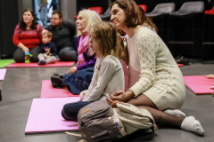 Uczestnicy zajęć siedzą na kolorowych matach. Na pierwszym planie kobieta w jasnej wełnianej sukience i dziecko przed nią. Obok leżący plecak.