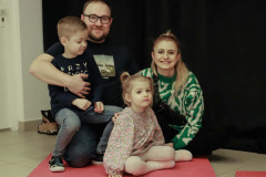 Na różowej macie siedzi rodzina. Mężczyna ma na sobie ciemną koszulkę z nadrukiem Babiego lata Chełmońskiego. Trzyma na kolanach chłopca. Uśmiechnięta kobieta ubrana w sweter w biało-zielone wzory. Przed nią dziewczynka w sukience w kwiatowy wzorek.
