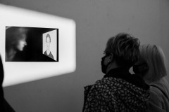 Dwie kobiety stoją tyłem do obiektywu. Oglądają oświetlone zdjęcie umieszczone na ścianie. Przedstawia ono zamglony profil mężczyzny wpatrzonego w popiersie wyświetlone na slajdzie. Zdjęcie czarno-białe.