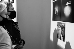 Dwie kobiety stoją bokiem do obiektywu. Wpatrują się w fotografie przymocowane do ściany. Zdjęcie czarno-białe.