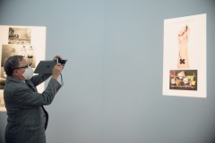 Mężczyzna w szarej marynarce fotografuje przymocowane do ściany zdjęcie. Zdjęcie przedstawia przedramię z naklejonym czarnym iksem.