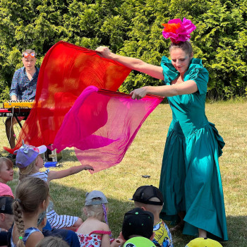 Kobieta w zielonej sukience z kwiatem na czubku głowy wymachuje płachtami materiału w kolorze różowym i czerwonym. Materiał próbują złapać dzieci. z