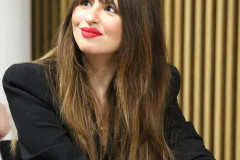 Zdjęcie portretowe. Anna Gacek trzyma długopis, patrzy w górę i uśmiecha się. Mocnym akcentem są usta pomalowane czerwoną szminką.