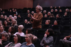 Publiczność siedząca w fotelach. Kobieta w centrum kadru stoi. Dłonie uniesione na wysokość piersi.