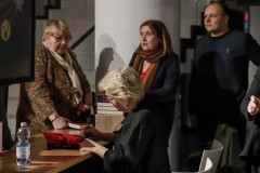 Maria Peszek w trakcie podpisywania ksiązek. Przy stoliku stoją trzy osoby. Artystka sfotografowana z lewego profilu.