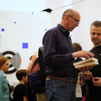 Prawa strona kadru Andrzej Dragan w czerni z długopisem. Obok niego widziany z prawego profilu mężczyzna w okularach, granatowej koszuli i dżinsach z dwiema książkami. Za nimi uczestnicy spotkania.