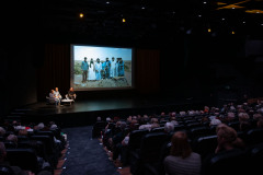 Zdjęcie zza rzędów. Na scenie trzy osoby przy stoliku. Wyświetlony slajd ze zdjęciem z podróży: mężczyźni w rzędzie ubrani w galabije.