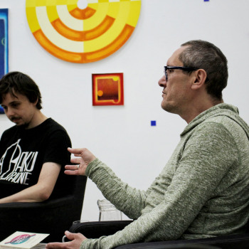 Sfotografowany z lewego profilu Robert Brzęcki siedzi w ciemnym fotelu. Za nim Piotr Tkacz z pochyloną głowa w ciemnej koszulce z białymi napisami. W głębi na białej ścianie wyeksponowane prace.
